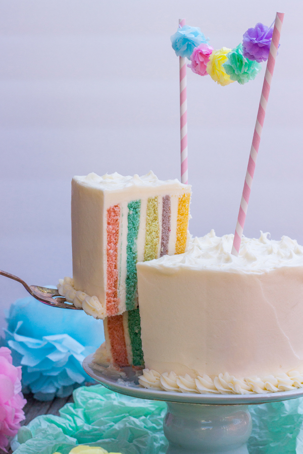 Pastel arcoiris en capas verticales recién partido, se ve la rebana de pastel siendo levantada del plato por una palita de plata