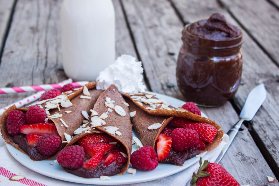 tres crepas de chocolate rellenas de frutos rojos en un plato, al fondo se ve un frasco con nutella y un vaso de leche