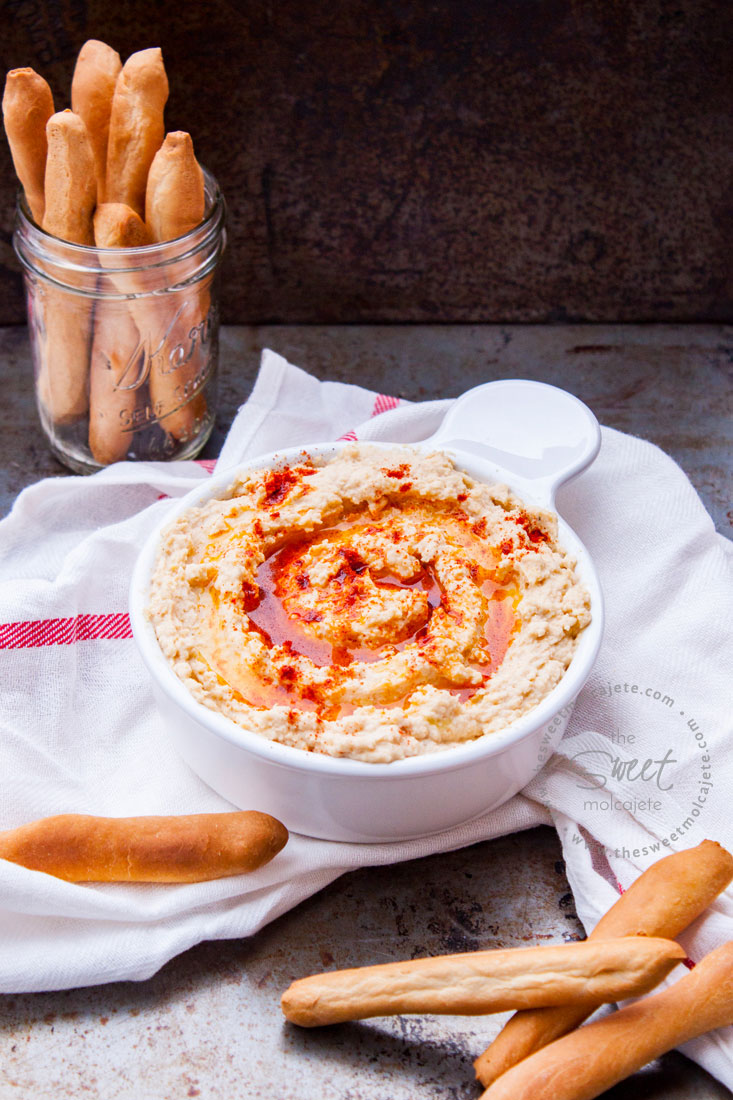 Receta básica de hummus de garbanzo dentro de un recipiente de cerámica blanco y acompañado de palitos de pan.