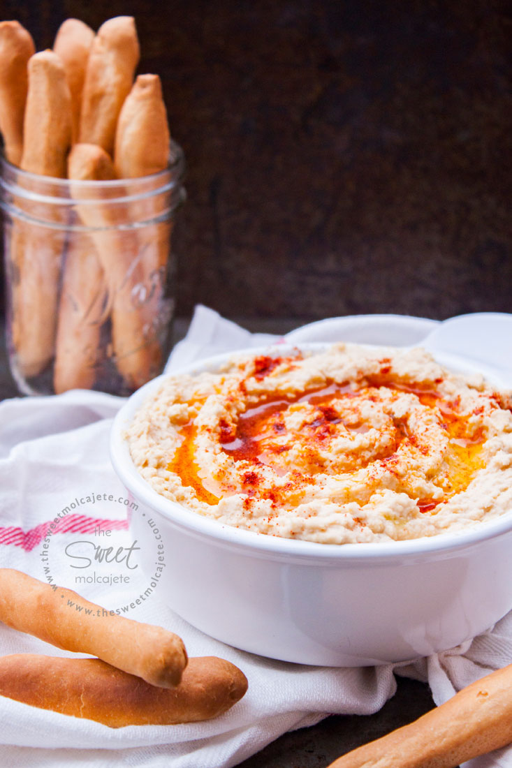 Receta básica de hummus de garbanzo dentro de un recipiente de cerámica blanco y acompañado de palitos de pan.