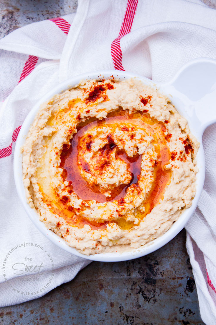 Vista aérea de un recipiente con Hummus con aceite de oliva y paprika en la superficie. El recipiente descansa sobre una servilleta de tela blanca.