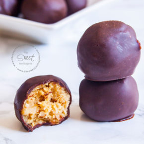 2 Bolitas de Cacahuate con chocolate una sobre otra y otra bolita mordida a un lado. Al fondo se ve un plato lleno de bolitas energéticas de cacahuate con chocolate