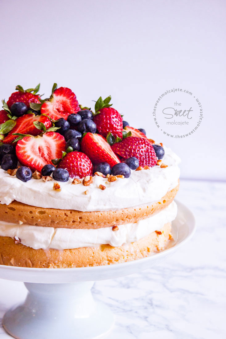 Pastel de tres leches en un pedestal de cerámica, el pastel está cubierto con crema batida, fresas, moras y nuez